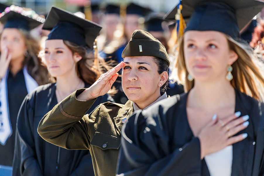 Graduation ceremony.  one graduate is in military fatigue saluting and the others are in traditional black graduation gowns and caps with their hands over their hearts.